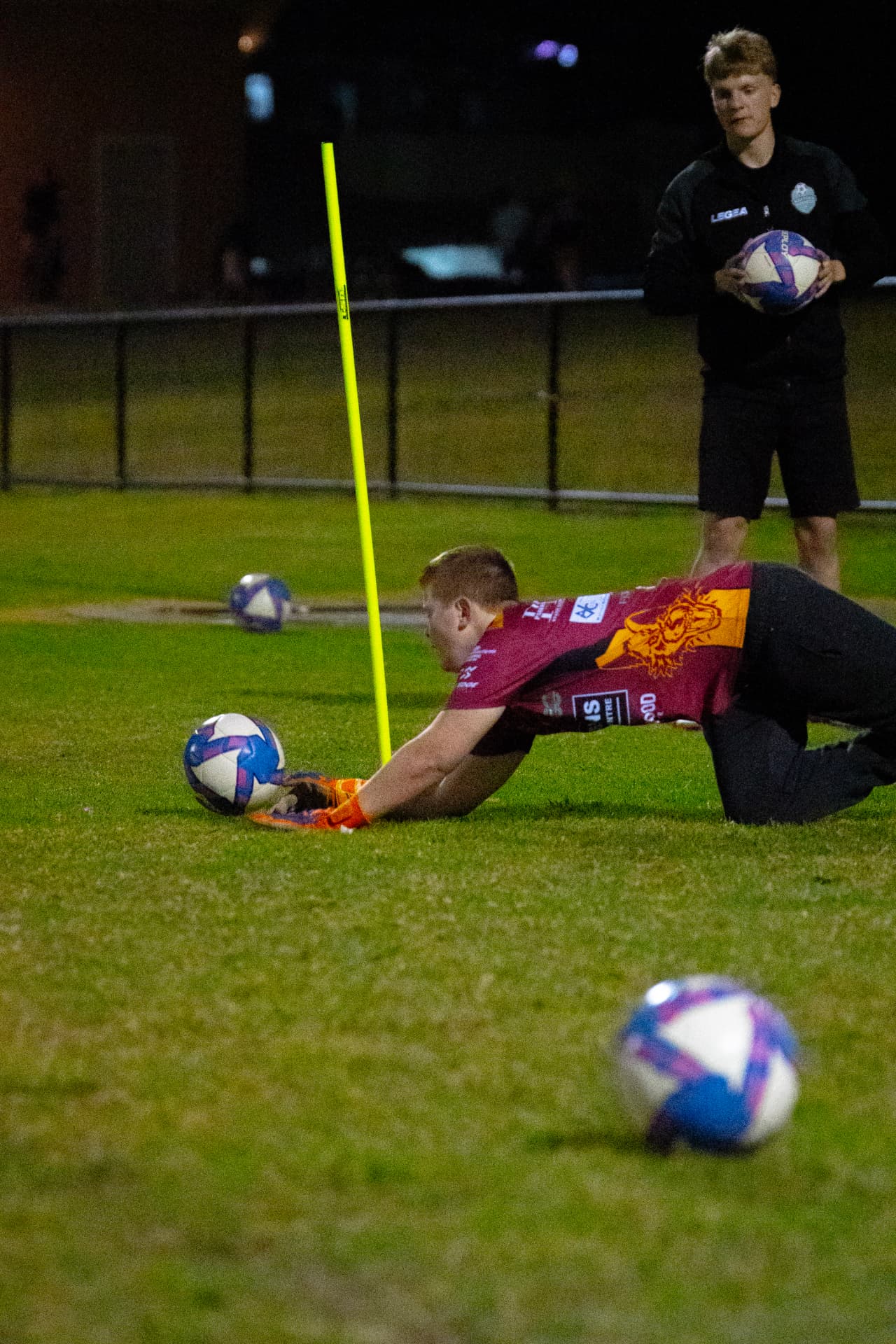 Goalkeeper in a maroon kit diving to smother a low ball beside a coach