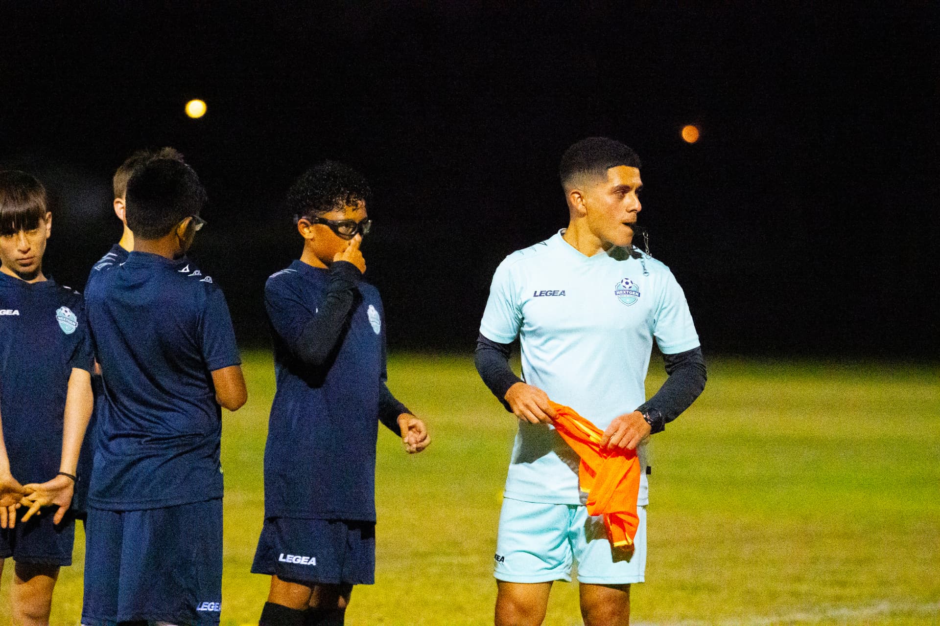 NextGen coach blowing the whistle beside players in navy kits