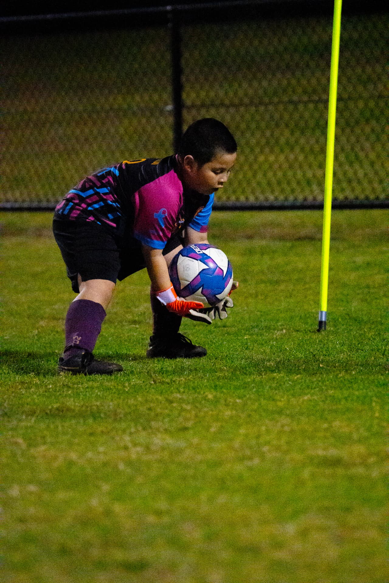 Young goalkeeper in orange gloves gathering the ball beside a training pole