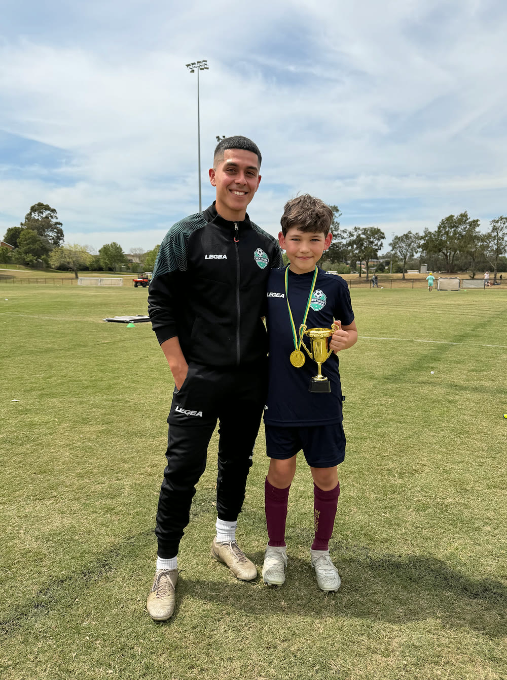 NextGen coach standing with a young player who is holding a trophy and medals